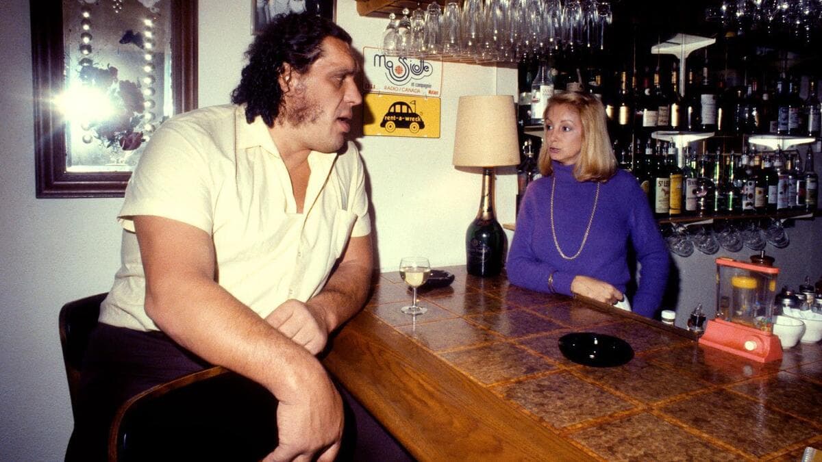 Andre the Giant with a glass of wine in front of him chatting to a blonde woman in purple attire