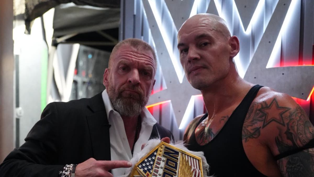 Baron Corbin holding up the WWE United States Title as Triple H points at him
