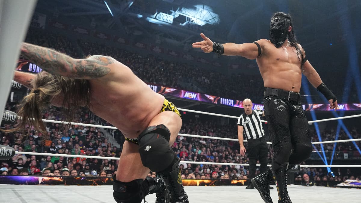 Chris jericho slumped on wrestling ring ropes as Bandido in his lucha mask points a finger gun at his back.