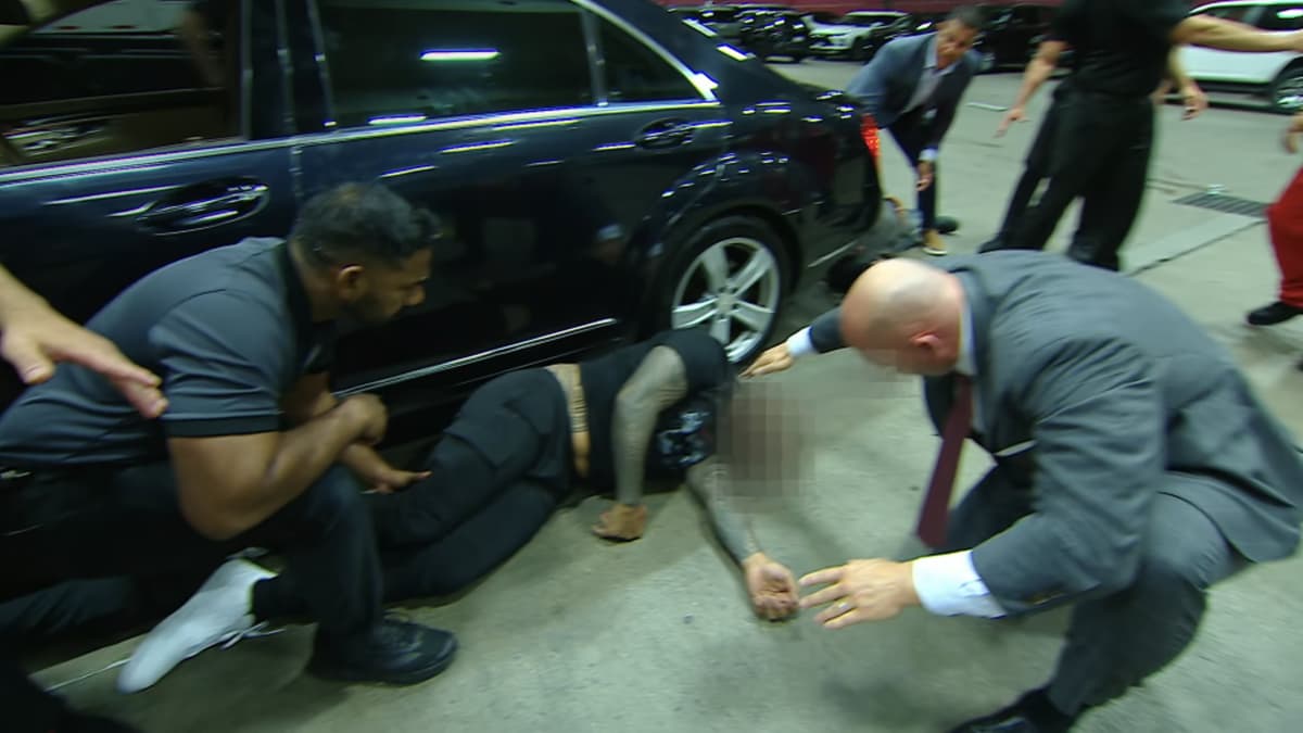Jey Uso being checked on by Adam Pearce while lay next to a car with a blurred face