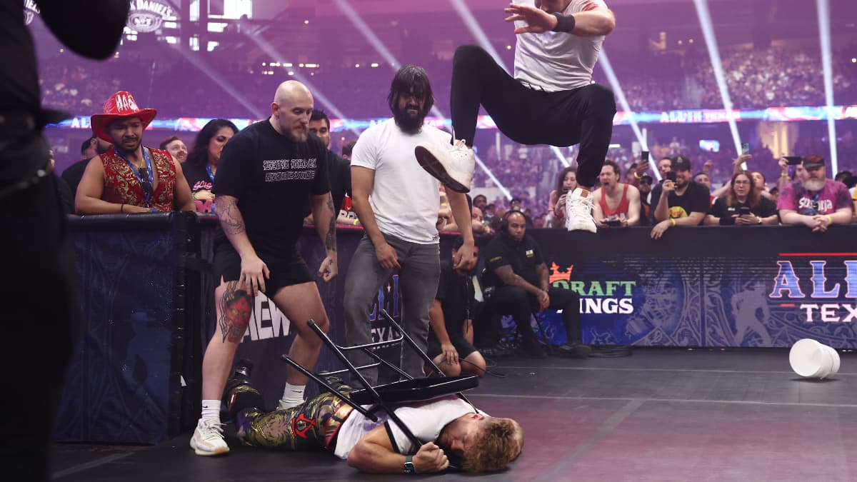 Claudio Castagnoli leaps high to stomp a chair wrapped around a downed Will Ospreay's neck, as Wheeler Yuta, Gabe Kidd, and an audience watch on