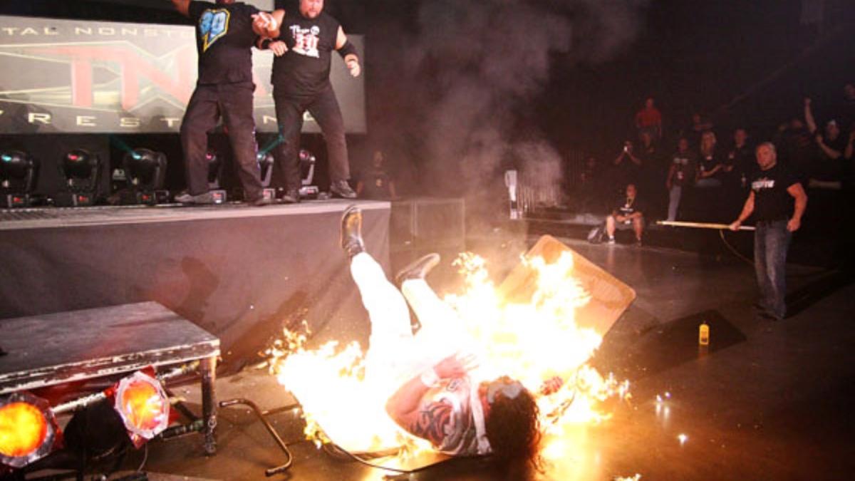 Abyss lying on a flaming table as Team 3D watch from the stage