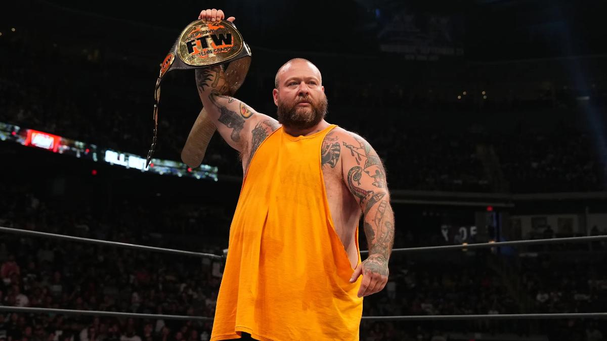 Action Bronson frowning in a sleeveless yello top holds up the FTW Championship in an AEW ring