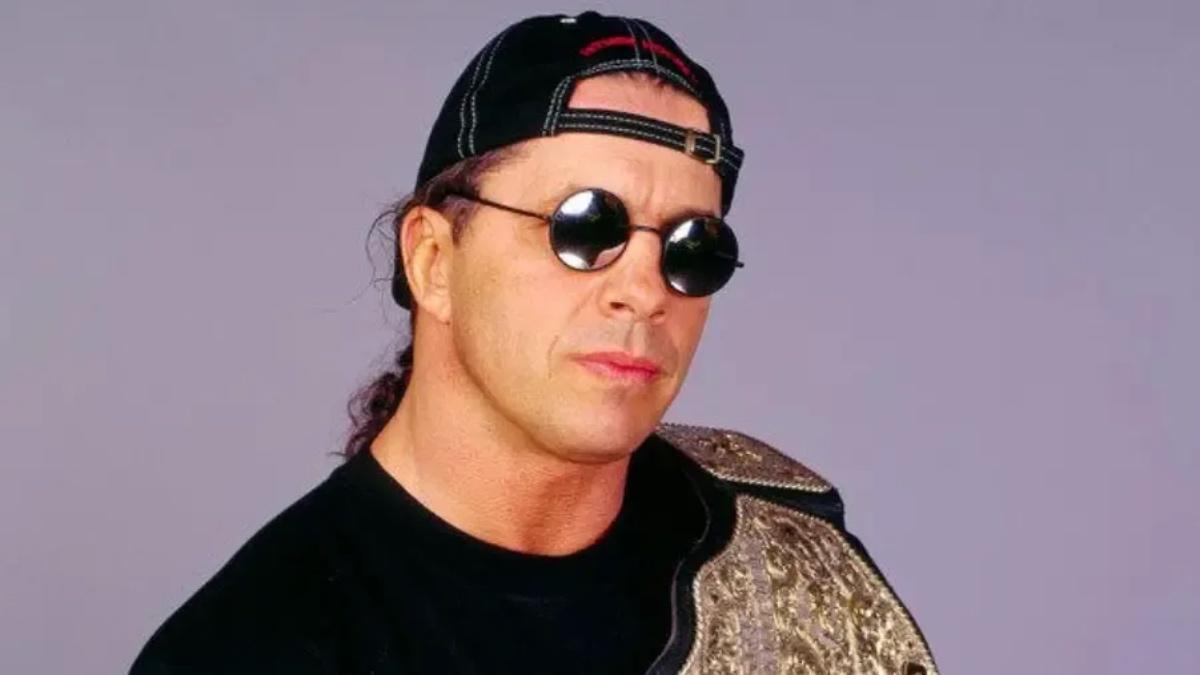 Bret Hart wearing black round sunglasses and a baseball cap backwards with the WCW World Heavyweight Title draped over his shoulder