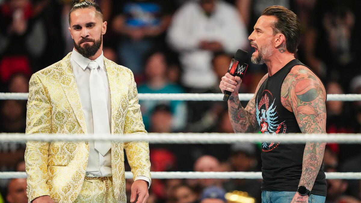 Seth Rollins in a gold suit looks on as CM Punk yells at him in a wrestling ring