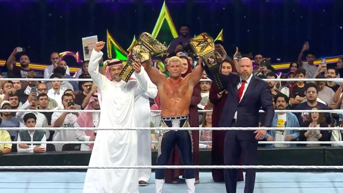 Cody Rhodes holds up two championship belts in a wrestling ring flanked by officials and Triple H