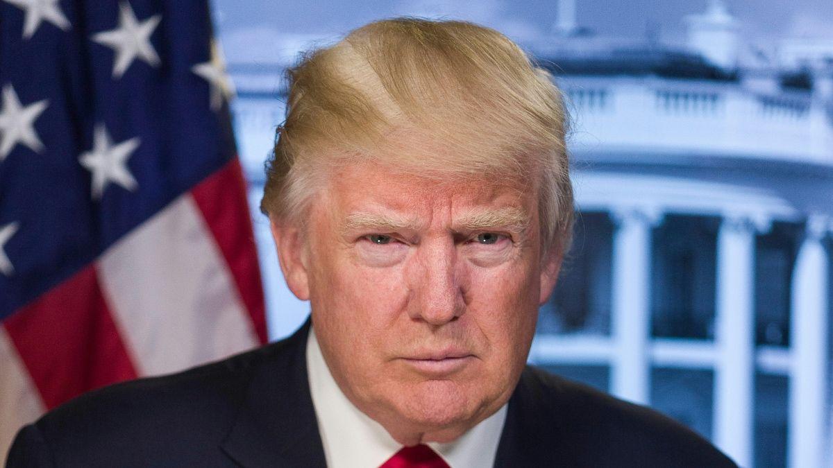 Donald Trump sitting in front of an American flag in a suit with a red tie