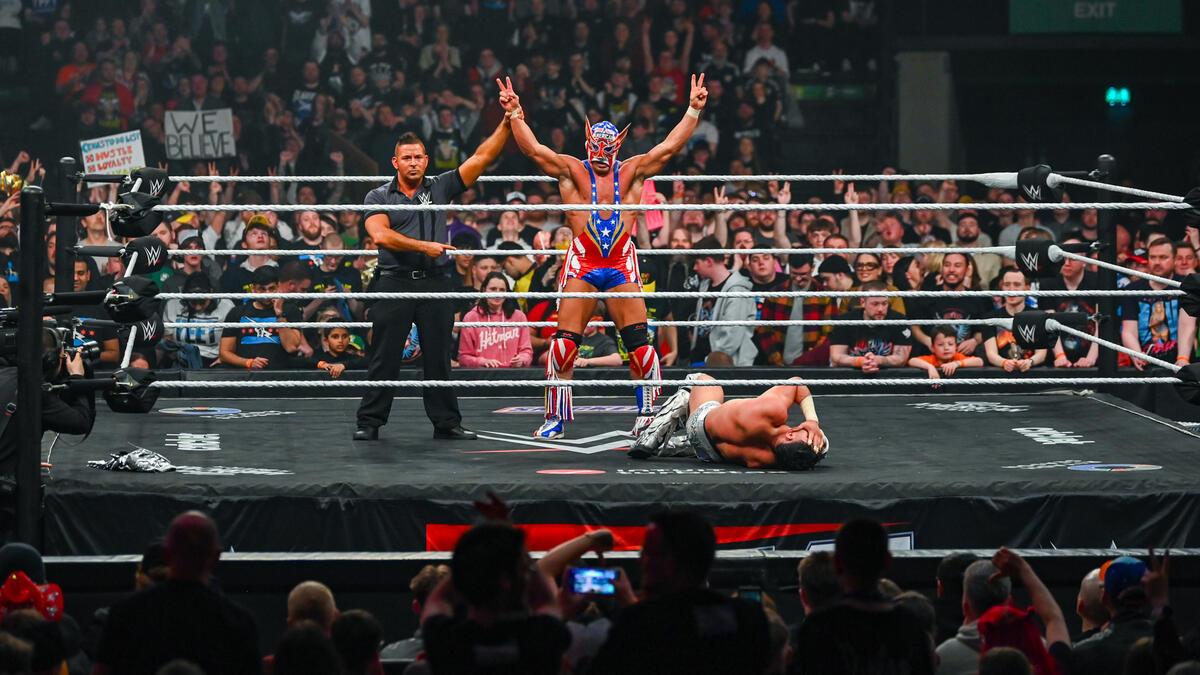 El Grande Americano has his hands raised by the referee in a wrestling ring after winning a match, whilst his opponent lays on the ring mat.