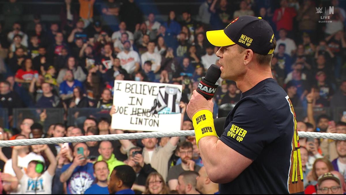 John Cena cutting a promo during WWE Raw while an 'I Believe In Joelinton' sign appears behind him 