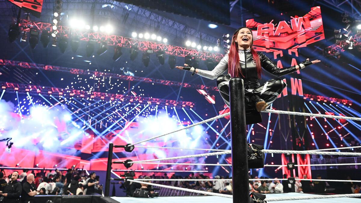 IYO SKY sits cross legged and smiling on a wrestling ring turnbuckle as fireworks go off in the background