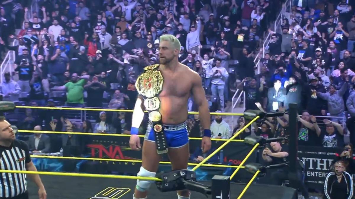 Joe Hendry stands on the middle turnbuckle in a TNA wrestling ring with the TNA World Championship belt over his shoulder