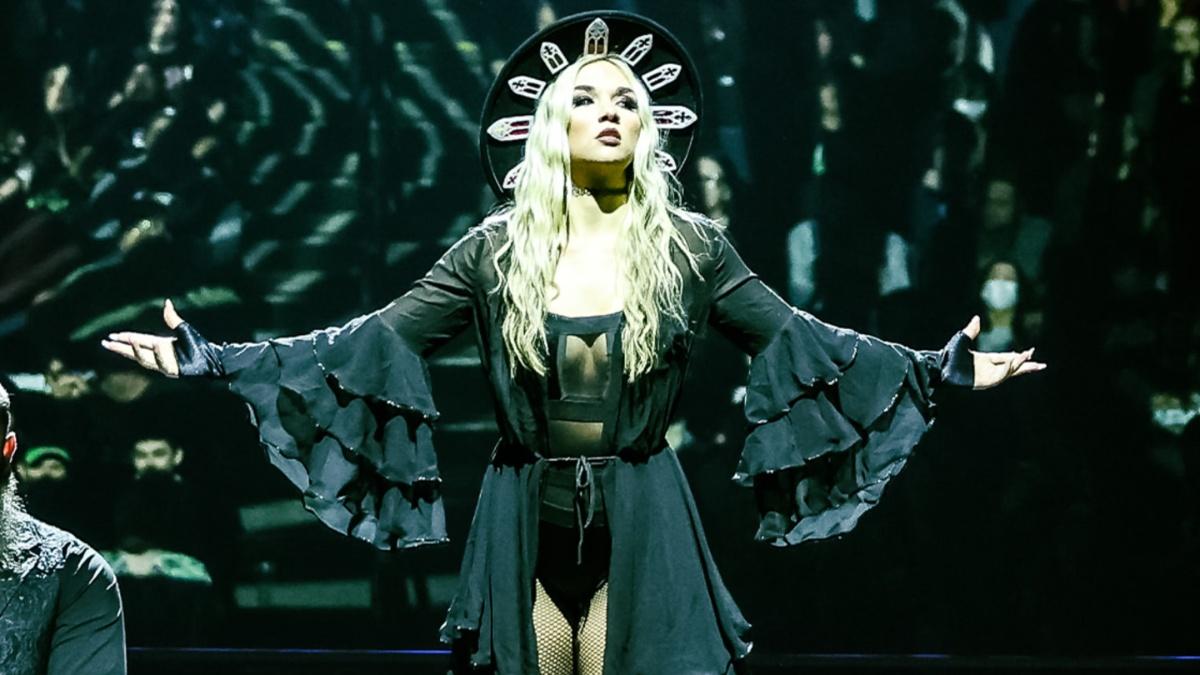 Julia Hart in a black outfit with dramatic sleeves and a headpiece, arms extended on stage.