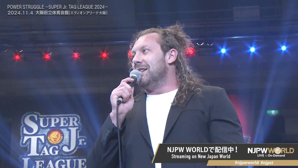 Kenny Omega in a suit jacket and white t-shirt hodling a microphone in an arena