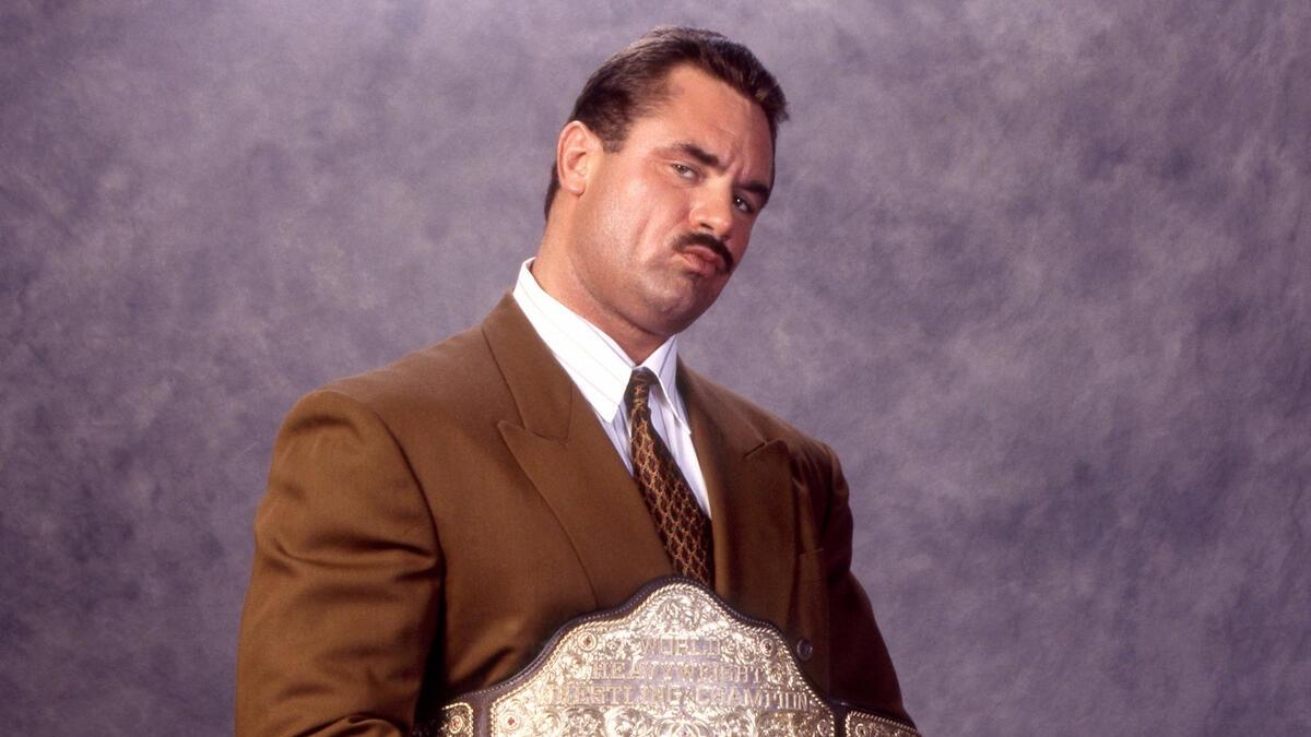 Rick Rude, wearing a lovely brown suit, poses for a promotional photo with the WCW World Heavyweight Championship belt