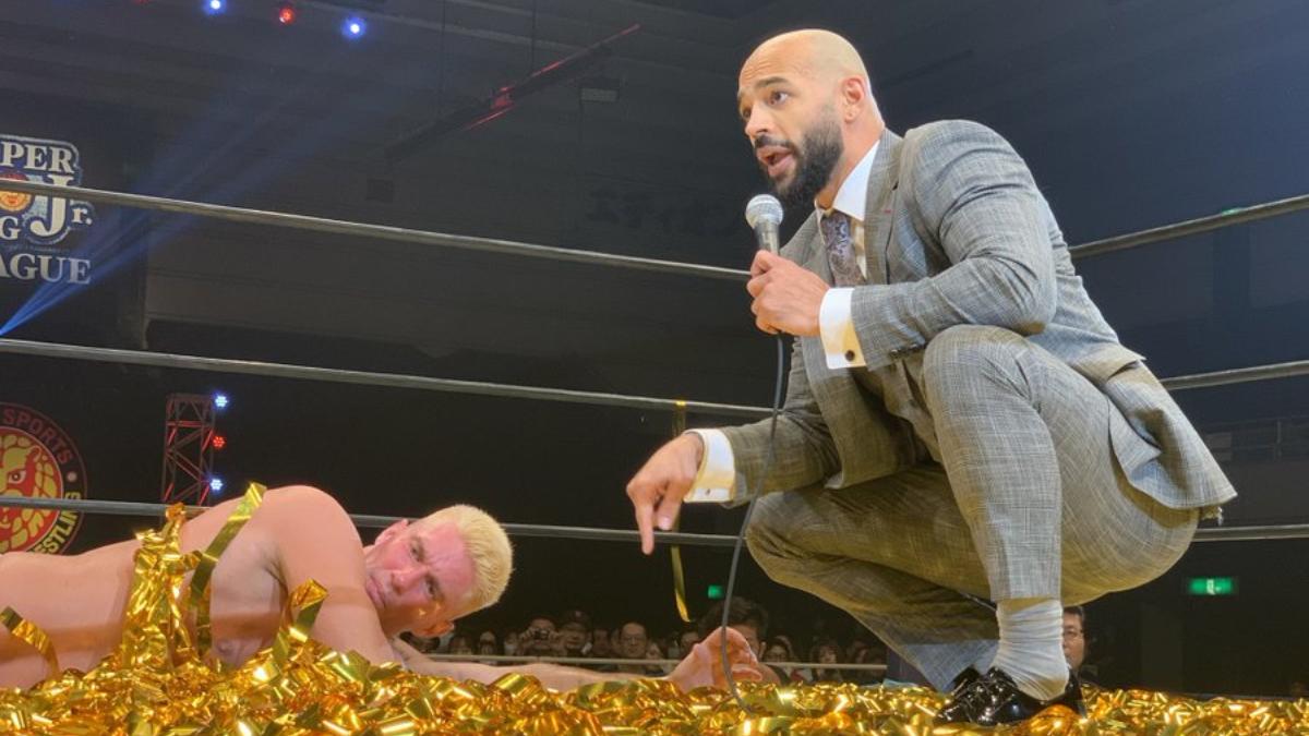 Ricochet crouching in a suit pointing at Zack Sabre Jr. lying on gold streamers in a wrestling ring.