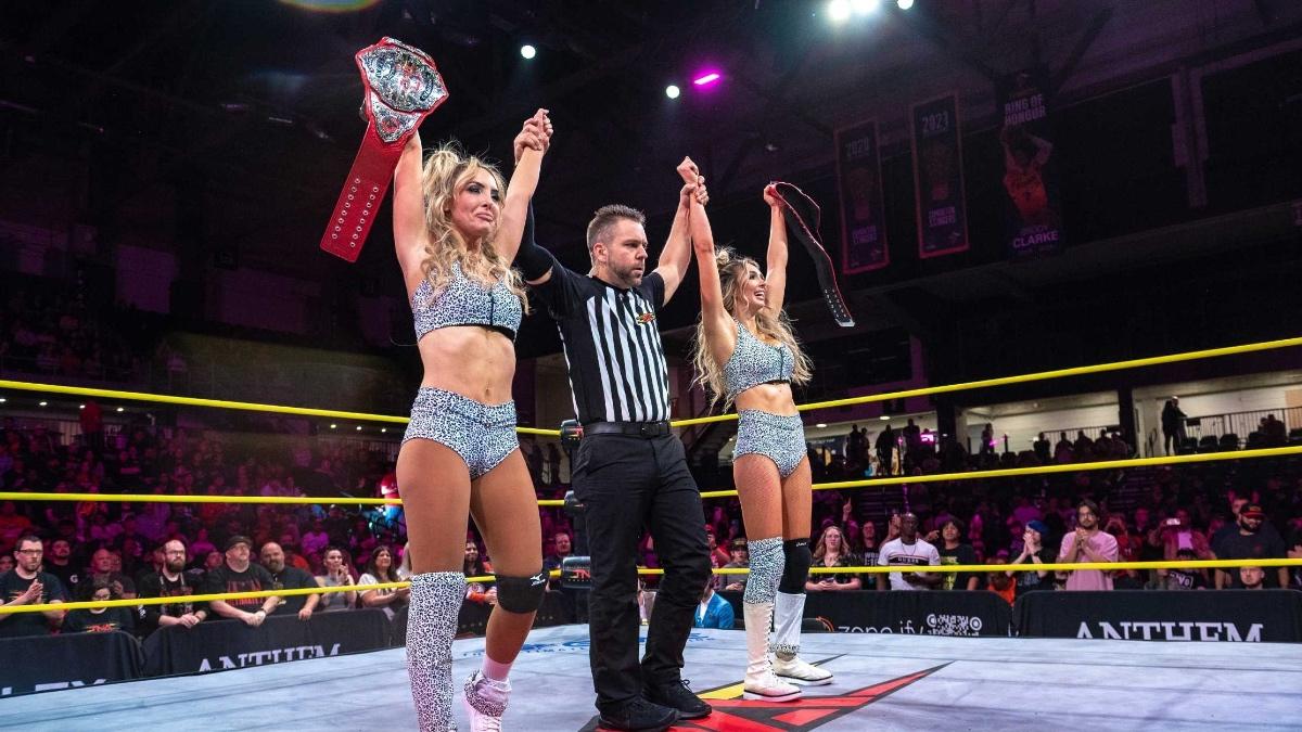 The IInspiration holding up the TNA Knockouts World Tag Team Titles with their arms raised by the referee, who is stood in the middle of them inside a pro wrestling ring
