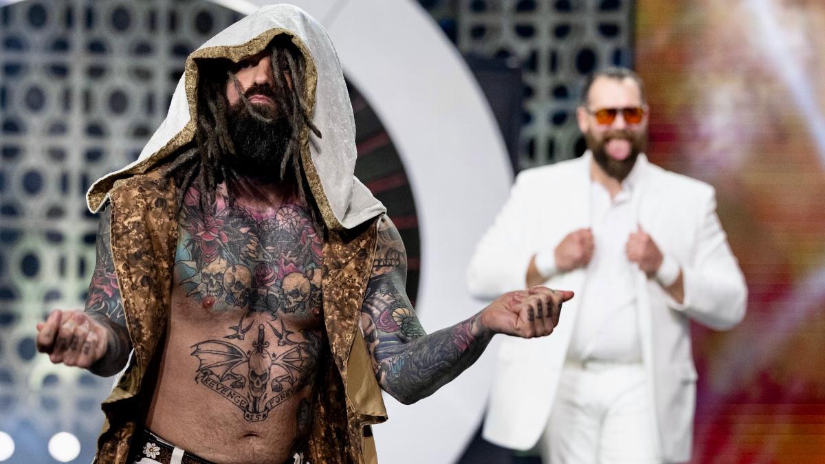 Vincent in a cape walks to  a wrestling ring, with Dutch obscured in the background in a white suit and sunglasses.