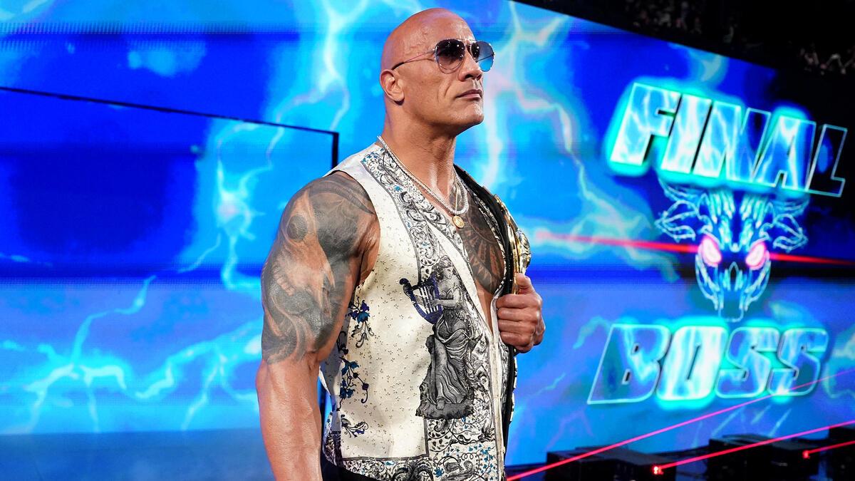 The Rock in a cream vest standing on the entrance way wearing sunglasses and holding his People's Champion title belt over his left shoulder