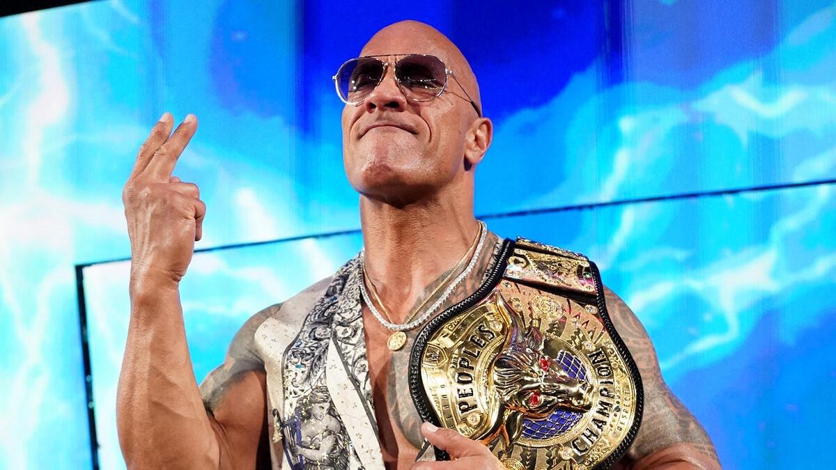 The Rock with his People's Champion belt in a vest holding up two fingers 