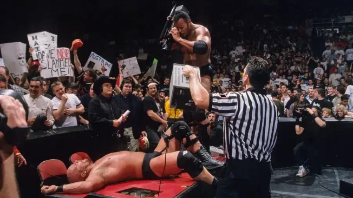 The Rock pointing a camera at Steve Austin while Austin is sprawled across the announce table at WWE Backlash 1999