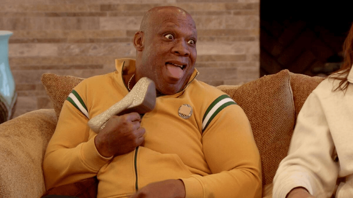 Tony Atlas holds a woman's shoe and sticks his tongue out during an episode of WWE Legends House