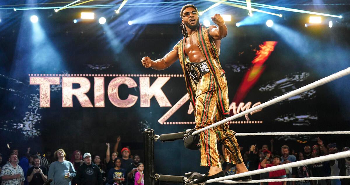 Trick Williams in a gold outfit with the NXT Title belt around his waist standing on the middle turnbuckle