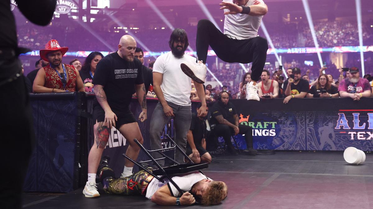 Claudio Castagnoli leaps high to stomp a chair wrapped around a downed Will Ospreay's neck, as Wheeler Yuta, Gabe Kidd, and an audience watch on