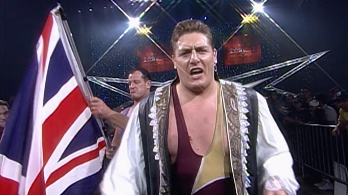 William Regal in a singlet looking at the camera as Dave Taylor waves the Union flag