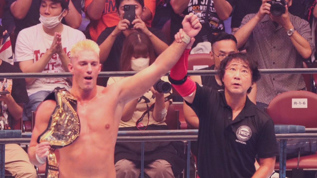 Zack Sabre Jr. with his arm raised by Red Shoes Uno with the IWGP World Heavyweight Title belt across his right shoulder 